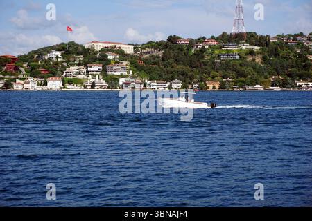 Istanbul, Türkei. Die größte Stadt in der Türkei und eine der wenigen transkontinentalen Städte der Welt, die sich über Europa und Asien auf der Bosporus-Straße erstreckt. Ein kleines weißes Motorboot mit rot-weiß gestreifter Markise, das auf einem tiefblauen Gewässer segelt. Das Boot bewegt sich in Richtung der rechten Seite des Bildes und hinterlässt eine weiße Spur. Im Hintergrund ist ein Hügel mit üppigen grünen Bäumen und Häusern zu sehen, mit einer roten Flagge, die auf einem der Gebäude thront. Ein hoher, weißer Turm mit einer rot-weiß gestreiften Spitze ist ebenfalls auf dem Hügel zu sehen. Der Himmel ist ein klares Blau mit ein paar verstreut Stockfoto