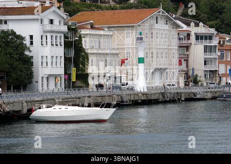 Istanbul, Türkei. Die größte Stadt in der Türkei und eine der wenigen transkontinentalen Städte der Welt, die sich über Europa und Asien erstreckt und über den Bosporus verläuft, Eine ruhige Hafenszene. Ein weißes Segelboot mit rotem Streifen ist an einem Betonsteg angedockt, der von einem Metallgeländer begrenzt wird. Der Pier befindet sich neben einer Reihe weißer Gebäude mit roten Dächern, von denen einige Balkone haben. Ein weißer Leuchtturm mit grüner Spitze steht gut sichtbar auf dem Pier. Das ruhige Wasser reflektiert die Gebäude und den Leuchtturm und schafft eine ruhige Atmosphäre. Einige Leute können gesehen werden, wie sie entlang des Piers spazieren gehen, und ein kleines Boot Stockfoto