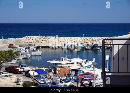 Latsi (Latchi), Zypern. Die Nordwestküste Zyperns im Bezirk Paphos, nahe der Stadt Polis. Entspanntes, malerisches Dorf am Meer, bekannt für seinen Yachthafen, frische Meeresfrüchte und ruhige Strände. Eine ruhige Hafenlandschaft mit verschiedenen Booten, die im ruhigen, blauen Wasser anlegen. Der Hafen liegt an einem felsigen Wellenbrecher, mit dem tiefblauen Ozean, der sich bis zum Horizont erstreckt. Der Himmel ist klar, hellblau, und das Wasser scheint relativ ruhig. Der Hafen ist von einer Steinmauer umgeben und einige Autos parken am Ufer. Ein kleines Boot mit blauem Baldachin liegt in der Nähe des an Stockfoto