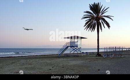 Zypern, Larnaka, Eine ruhige Strandszene bei Sonnenuntergang. Der Himmel ist in Orange- und Rosa-Tönen gemalt und bildet eine wunderschöne Kulisse für die untergehende Sonne. Der Ozean, ein tiefes Blau, erstreckt sich bis zum Horizont, mit kleinen Wellen, die sanft über die Küste schlagen. Im Vordergrund steht ein Rettungsschwimmer mit rotem Dach und blauem Sockel am Sandstrand, begleitet von einer einsamen Palme. Ein weißes Flugzeug wird während des Fluges gefangen genommen und bewegt sich von links nach rechts über den Himmel. Einige Liegen sind auch in der Nähe des Rettungsschirms zu sehen. Das Auge und die Phantasie eines Künstlers Stockfoto