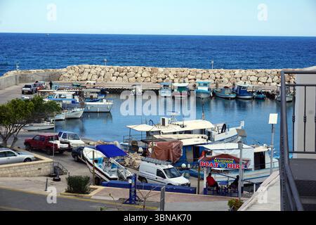 Latsi (Latchi), Zypern. Die Nordwestküste Zyperns im Bezirk Paphos, nahe der Stadt Polis. Entspanntes, malerisches Dorf am Meer, bekannt für seinen Yachthafen, frische Meeresfrüchte und ruhige Strände. Eine Hafenszene mit einer Vielzahl von Booten und Yachten im ruhigen, blauen Wasser. Der Hafen ist von einer Steinmauer und einem Betonpfeiler umgeben, auf dem einige Personen zu sehen sind. Der Himmel ist klar, hellblau, und das Wasser ist relativ ruhig. Die Boote sind meist weiß und blau, mit einigen rot-weißen Booten. Ein kleiner weißer Van parkt in der Nähe des Piers. Ein Schild für „Bootsverleih“ ist sichtbar Stockfoto