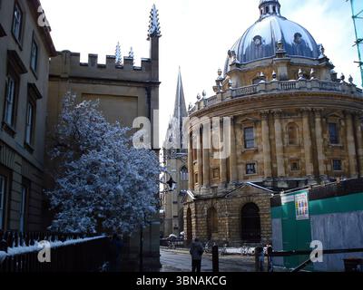 Radcliffe Camera, Bodleian Library, Oxford – die berühmte Stadt in England, die weltweit für ihre renommierte Universität und reiche Geschichte bekannt ist, Großbritannien. Eine ruhige Winterszene mit einer renommierten Bibliothek und Forschungszentrum. Das Gebäude mit seiner markanten Kuppel und seinen komplexen architektonischen Details steht majestätisch im Hintergrund. Im Vordergrund fügt ein Baum mit schneebedeckten Ästen der Stadtlandschaft einen Hauch von Natur hinzu. Ein schwarzer Zaun mit einer schneebedeckten Spitze verläuft entlang der linken Seite des Bildes, und ein Teil eines Gebäudes mit einem zinnenförmigen Turm ist links zu sehen. Auf dem Stockfoto