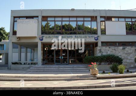Ein modernes zweistöckiges Gebäude mit einer hellen beigefarbenen oder cremefarbenen Fassade. Das Gebäude verfügt über große Fenster im zweiten Stock und einen kleineren Abschnitt auf der linken Seite. Der Eingang ist durch eine Treppe gekennzeichnet, die zum Haupteingang führt, der von einer Glastür eingerahmt ist. Der Name des Gebäudes wird über dem Eingang in einem blauen Schild angezeigt. Das Gebäude liegt an einer Straße mit einem gepflasterten Bürgersteig und einem kleinen Blumenbeet davor. Ein kleiner Teil eines zweiten Gebäudes ist links vom Hauptgebäude zu sehen. Der Himmel ist klar blau und Bäume sind im Hintergrund zu sehen. Ein AR Stockfoto