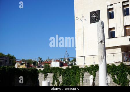 Eine hohe, weiße Steinsäule mit einem sichtbaren Riss oder einer Beschädigung auf der rechten Seite, die in einem Innenhof steht. Die Säule ist von einer niedrigen Steinmauer mit grünem Efeu umgeben. Im Hintergrund ist ein modernes Gebäude mit einer Uhr an der Fassade sowie ein Teil eines Gebäudes mit kuppelförmiger Struktur zu sehen. Der Himmel ist klar, leuchtend blau. Der Innenhof ist von einem Metallgeländer begrenzt, und in der Ferne sind einige Personen zu sehen. Plovdiv, Bulgariens zweitgrößte Stadt, ist eine der ältesten, ständig bewohnten Städte Europas mit einer Geschichte Stockfoto
