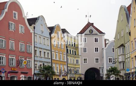 Schärding, das charmante Barockjuwel am Inn in Oberösterreich. Bekannt für die farbenfrohe barocke Silberzeile mit pastellfarbenen Zunfthäusern und gut erhaltenen mittelalterlichen Mauern und Toren. Eine Straßenszene mit einer Reihe von bunten Gebäuden mit Giebeldächern und weißen Zierleisten. Die Gebäude sind in Rosa-, Gelb-, Blau- und Grüntönen gestrichen und etwas linear angeordnet. Ein markantes Merkmal ist ein großes, helles Gebäude mit einem Uhrturm und einer Wetterfahne auf der Spitze. Die Straße ist gesäumt von geparkten Autos, und einige Leute können auf dem Gehweg gesehen werden. Der Himmel ist Stockfoto