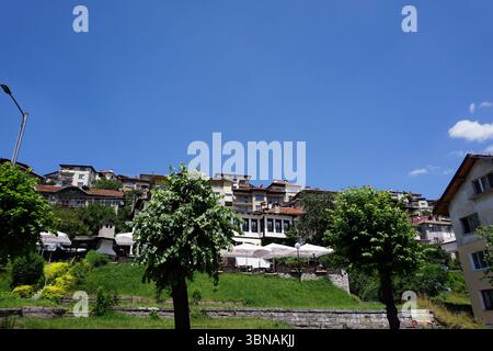 Smolyan, Bulgarien, Eine malerische Stadt am Hügel mit einer Ansammlung von Gebäuden im traditionellen Architekturstil, mit roten Ziegeldächern und weißen Wänden. Die Gebäude sind terrassenförmig oder stufenförmig angeordnet, einige Gebäude haben Balkone. Die Stadt liegt eingebettet in üppiges Grün mit Bäumen und Büschen, die die Landschaft prägen. Mehrere weiße Sonnenschirme stehen auf einem grasbewachsenen Bereich vor den Gebäuden, die auf Sitzgelegenheiten oder Essbereiche im Freien hinweisen. Eine Steinmauer oder Stützkonstruktion ist im Vordergrund sichtbar, und eine Straßenlaterne ist auf der linken Seite des Bildes vorhanden. Klar Stockfoto