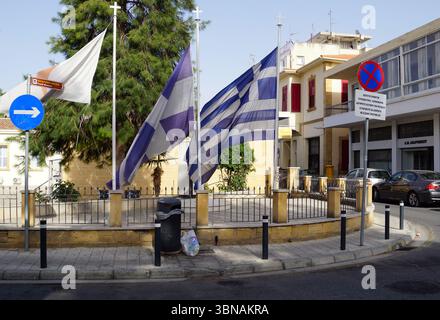 Nikosia (Lefkosia), Zypern, die letzte geteilte Hauptstadt der Welt, Eine Straßenecke, an der drei Flaggen an den Fahnenmasten deutlich zu sehen sind. Eine Flagge mit ihren markanten blauen, weißen und lila Farben befindet sich links, eine weiße und blaue Flagge rechts. Eine weiße und blaue Flagge mit einem weißen Pfeil ist ebenfalls auf der linken Seite sichtbar. Die Fahnen flattern im Wind und verleihen der Szene ein Gefühl von Bewegung. Die Fahnen sind an Fahnenmasten befestigt, die sich auf einer niedrigen Steinmauer mit einem schwarzen Metallzaun befinden. Ein schwarzer Mülleimer befindet sich in der Nähe der Wand. Im Hintergrund ein Gebäude Stockfoto