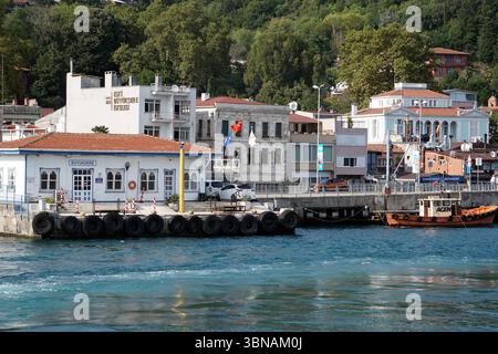 Istanbul, Türkei. Die größte Stadt in der Türkei und eine der wenigen transkontinentalen Städte der Welt, die sich über Europa und Asien auf der anderen Seite des Bosporus erstreckt. Eine ruhige Hafenszene mit einem weißen Gebäude mit einem roten Dach und einem blauen Schild mit der Aufschrift „BUYKUDE“ auf der linken Seite. Das Gebäude befindet sich auf einem Betonpier, in der Nähe liegt ein kleines Boot. Der Pier ist mit mehreren schwarzen Reifen ausgekleidet, und ein kleines Dock erstreckt sich davon. Ein rot-weißes Boot legt am Pier an. Im Hintergrund liegt eine Reihe von Gebäuden mit roten Dächern und weißen Wänden, einige mit Balkonen, inmitten von üppigem Grün Stockfoto
