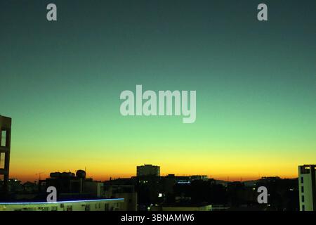 Larnaka, Zypern, das Bild fängt eine Skyline bei Sonnenuntergang ein, mit einem verlaufenden Himmel, der von Dunkelblau zu einer lebhaften Mischung aus Grün-, Gelb- und Orangetönen übergeht. Die Skyline ist im Hintergrund sichtbar, mit Gebäuden in unterschiedlicher Höhe und Farbe, darunter auch mit reflektierenden Oberflächen. Der Himmel ist größtenteils klar, mit ein paar schwachen Wolken. Das Bild wird von einem hohen Aussichtspunkt aufgenommen und bietet einen Panoramablick auf die Skyline und den Himmel. Die Gebäude sind größtenteils gegen den farbenfrohen Sonnenuntergang gehalten, mit ein paar schwachen Lichtern, die in der Entfernung sichtbar sind. Bildunterschrift in Form eines Künstlerauges und einer phantasievollen Bildunterschrift. Stockfoto