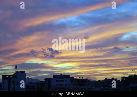 Larnaka, Zypern, das Bild fängt eine Skyline bei Sonnenuntergang ein, mit einem leuchtenden Himmel voller Orange-, Gelb- und Blautöne. Der Himmel ist mit Wolken gespickt, was der Szene Tiefe und Textur verleiht. Das Stadtbild umfasst eine Vielzahl von Gebäuden, darunter Wohn- und Geschäftsgebäude, mit einigen Dächern, die mit Antennen und Satellitenschüsseln verziert sind. Die Silhouetten der Gebäude vor dem farbenfrohen Himmel bilden einen beeindruckenden Kontrast. Das Stadtbild liegt auf einem Hügel mit Bergen, die in der Ferne sichtbar sind. Das Bild wird aus einem hohen Blickwinkel aufgenommen und bietet einen umfassenden Überblick über Stockfoto