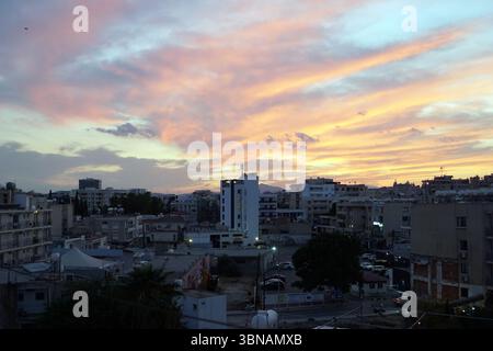 Larnaka, Zypern, das Bild fängt eine Stadtlandschaft bei Sonnenuntergang ein, mit einem leuchtenden Himmel voller rosa, orangen und gelben Tönen. Die Szene ist eine Mischung aus Gebäuden, darunter ein hohes weißes Gebäude mit einem blau/grauen Dach und eine Vielzahl anderer Strukturen in Beige-, Grau- und Brauntönen. Die Gebäude sind dicht gepackt, was ein Gefühl von städtischer Dichte schafft. Der Himmel ist eine wunderschöne Leinwand aus Farben, mit dem Sonnenuntergang hinter den Gebäuden, der ein warmes Licht über die Szene wirft. Das Bild wird aus einem hohen Blickwinkel aufgenommen und bietet einen Panoramablick. Der Vordergrund umfasst eine Mischung aus Bäumen und Vegetation, Stockfoto