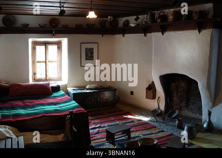 Bansko ist eine malerische Stadt im Südwesten Bulgariens, eingebettet am Fuße des Piringebirges. Ein gemütliches und rustikales Zimmer mit Kamin als zentrales Element. Der Kamin ist aus weißem Stuck gebaut und hat einen dunklen, möglicherweise Ziegelofen. Über dem Kamin befindet sich ein Holzregal mit verschiedenen Gegenständen, darunter mehrere dunkle Töpfe und Pfannen und einige dekorative Gegenstände. Der Raum wird durch eine einzelne Leuchte beleuchtet, die von der Decke hängt und ein warmes Licht auf die weißen Wände wirft. An einer Wand befindet sich ein Fenster mit Holzrahmen, sodass natürliches Licht in den Raum eindringen kann. Stockfoto
