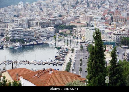 Kavala, Griechenland, das Bild zeigt einen Panoramablick auf einen Hügel mit Blick auf einen Hafen voller Boote und Yachten. Das Stadtbild ist dicht gefüllt mit Gebäuden in unterschiedlichen Höhen und Farben, hauptsächlich in weiß-, Beige- und Grautönen, mit einigen roten und orangen Akzenten. Der Hafen ist in leuchtendem Blau gehalten und mit Booten verschiedener Größen und Farben, darunter weiß, blau und rot, gespickt. Die Gegend ist von einer Mischung aus Bäumen und Vegetation umgeben, und der Himmel ist klar, hellblau. Eine Straße führt entlang des Ufers, an der entlang des Ufers ein Fußgängerweg oder eine Promenade zu sehen ist. Das Bild wird aufgenommen Stockfoto