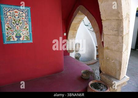 Eine leuchtend rote Wand mit blau-weißen Fliesen, die möglicherweise ein florales oder botanisches Muster darstellt. Die Mauer ist Teil eines überdachten Gehwegs oder einer Veranda, mit einem geschwungenen Bogen aus hellbeigem Stein, der die Aussicht umrahmt. Durch den Torbogen ist ein weißes Gebäude mit einem geschwungenen Dach sichtbar. Eine kleine Topfpflanze sitzt auf dem roten Boden im Vordergrund, und ein kleiner, dunkler Topf mit einer Pflanze befindet sich auf der rechten Seite des Gehwegs. Der Fußweg liegt auf einem hellen Fliesenboden. Zypern Dorf Stockfoto