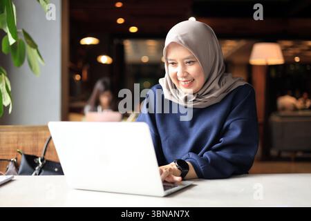 Muslimisch-asiatische Frauen mittleren Alters mit Hijab, die in einem Café arbeiten und einen Laptop benutzen. Stockfoto