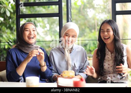 Drei asiatische Freundinnen, die glücklich im Café neben dem Fenster sitzen, lächeln, nachdem sie TikTok Challenge gemacht haben, zwei von ihnen tragen Hijab Stockfoto