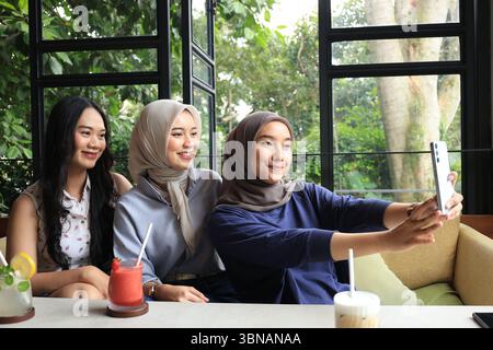 Drei asiatische Freundinnen glücklich sitzen im Café neben dem Fenster und nehmen Selfie auf dem Handy Stockfoto