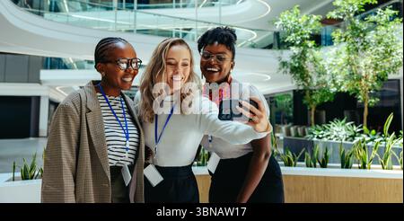 Drei fröhliche Geschäftsfrauen machen ein Selfie in einer hellen, modernen Büroumgebung. Das Bild spiegelt Themen wie Karrierefortschritt, Teamarbeit und c wider Stockfoto