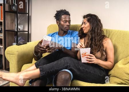 Ein liebevolles, interrassisches Paar teilt einen intimen Moment, indem er sich ansieht, während er zu Hause Popcorn isst. Stockfoto