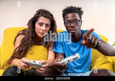 Ein junges Paar schaut und zeigt direkt auf die Kamera, während es ein Videospiel spielt. Stockfoto