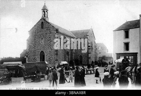 Ein belebter Dorfplatz in Rothéneuf liegt in einer Steinkirche mit Glockenturm und Bogenfenstern. Einheimische in historischen Kleidern versammeln sich an Marktständen und Wagen und erinnern an eine lebhafte Szene aus dem frühen 20. Jahrhundert. Die Architektur ist bescheiden, verwittert und deutlich bretonisch. Veröffentlicht in den 1910er Jahren von Léon & Lévy aus Paris. Von einer Originalpostkarte. Stockfoto