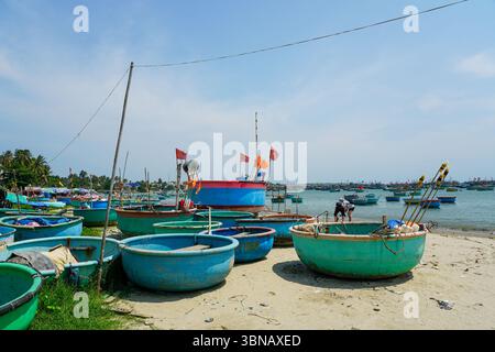 Mui ne, Vietnam – 21. März 2025: Traditionelle Rundkorbboote säumen den Sandstrand von Mui ne, mit fliegenden Fahnen und Fischerbooten auf dem Meer Stockfoto