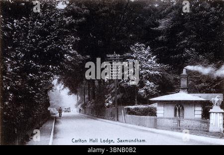 Eine ruhige, von Bäumen gesäumte Straße führt zur Carlton Hall Lodge in Saxmundham, deren Kamin sanft raucht. Ein rustikaler Zaun umgibt die Lodge, während ein Auto aus den frühen 1920er Jahren sich in der Ferne nähert – klein, aufrecht und erinnernd an eine neue Ära. Die Szene verbindet den edwardianischen Charme und moderne Verkehrsmittel. Von einer originalen, nicht zugeschriebenen Postkarte. Stockfoto