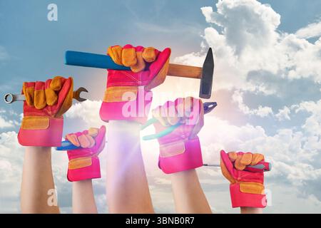 Die Hände des Mannes Arbeiter halten verschiedene Werkzeuge Hammer, Schraubendreher, Zange in Handschuhen vor blauem Himmel mit Wolken. Arbeitstag-Konzept. Stockfoto