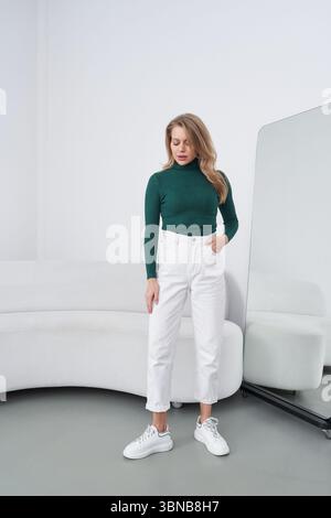 Lässiges Modebild, das eine Frau in grünem Rollkragenkragen und weißer Hose zeigt, neben einem Spiegel neben einem Sofa steht, weiße Sneakers trägt, mit Stockfoto