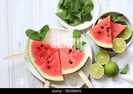 Wassermelonenscheiben, Limetten und Minze in weißen Platten, Draufsicht. Reife Wassermelonenstücke auf Stöcken, Holztisch und Limettenhälften. Sommeressen. Sommerfrüchte Stockfoto