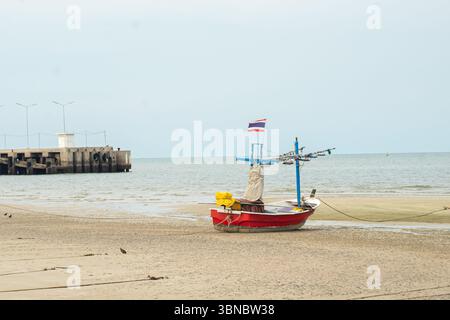 Ein kleines Fischerboot mit thailändischer Flagge an einem Sandstrand in der Nähe eines Piers unter einem trüben Himmel, Hua hin Resort, Thailand Stockfoto