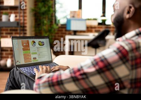 afroamerikaner, der zu Hause Marketingpläne analysiert und Geschäftsberichte erstellt. Schwarzer männlicher Unternehmer, der auf der Couch sitzt und sich auf die Projektpräsentation auf dem Laptop konzentriert. Stockfoto