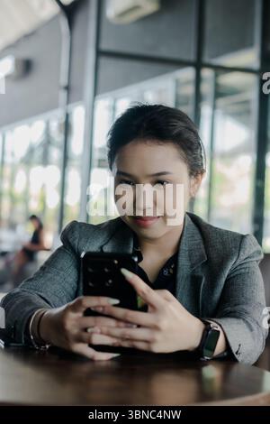 Eine elegante professionelle Frau macht eine Pause, um mit ihrem Smartphone zu interagieren. Sie sitzt an einem Tisch, wahrscheinlich in einem Café, mit einem ruhigen Ausdruck, Pos Stockfoto