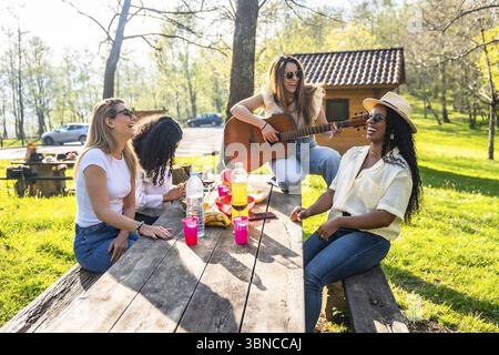 Vier Freundinnen sitzen an einem Holztisch, spielen Gitarre und genießen ein Picknick im Freien mit Getränken und Snacks Stockfoto