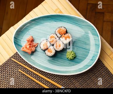 Frische Hosomaki-Brötchen, gefüllt mit Lachs, serviert mit eingelegtem Ingwer und Wasabi, präsentiert auf einem stilvollen Teller mit Essstäbchen in der Nähe Stockfoto
