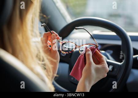Frau, die die Brille mit einem Tuch im Auto reinigt, bereitet sich auf ein sicheres und fokussiertes Fahren bei natürlichem Licht vor. Stockfoto