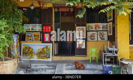 Farbenfrohe Kunstgalerie in der antiken Stadt Hoi an, Vietnam, mit einheimischer Frau und Hund vor einem traditionellen gelb bemalten Gebäude Stockfoto