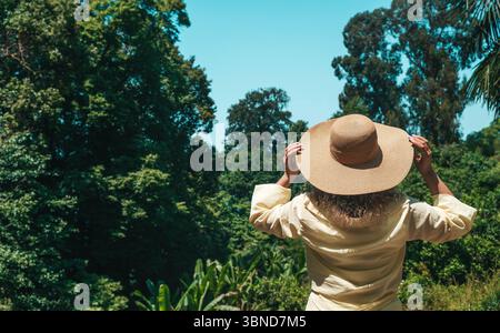 Rückansicht einer Frau, die ein hellgelbes Hemd und einen breitkrempigen Strohhut trägt und an einem sonnigen Tag vor einem üppigen tropischen Wald steht. Ihre Hände halten sanft den Hut, während sie auf die grüne Landschaft und den blauen Himmel blickt. Dieses Bild fängt einen ruhigen Moment des Reisens, der Naturwahrnehmung und der Achtsamkeit in einer natürlichen Umgebung ein, die perfekt für Themen wie langsames Leben, Ökotourismus und Sommertouren geeignet ist. Stockfoto