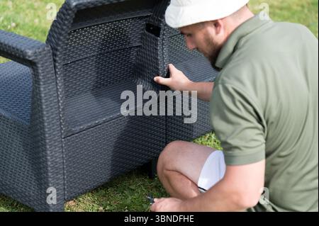 Ein Mann montiert dunkelgraue Korbmöbel auf einem grasbewachsenen Rasen. Das Packstück scheint Stauraum zu haben. Entspannen Sie sich und genießen Sie die Natur. Stockfoto