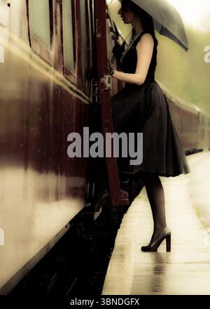 Eine Frau in schwarzem Kleid und Heels tritt auf einen Bahnsteig, während sie im Regen einen Regenschirm hält. Stockfoto