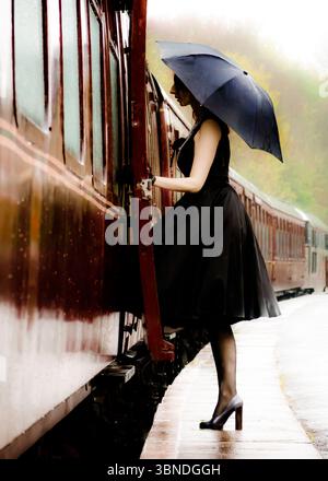 Eine Frau in schwarzem Kleid und Heels steigt in einen Zug, während sie an einem regnerischen Tag einen Regenschirm hält. Stockfoto