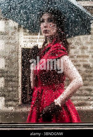 Eine Frau in rotem Kleid steht hinter einem regenbedeckten Fenster und hält einen Regenschirm. Stockfoto