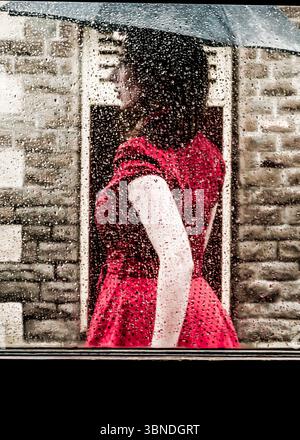 Eine Frau in rotem Kleid steht hinter einem Fenster, das mit Regentropfen bedeckt ist, und hält einen Regenschirm. Stockfoto