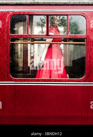 Eine Frau in einem roten Pünktchen-Kleid steht in einem Zugwagen, sichtbar durch ein regendurchflutetes Fenster. Stockfoto