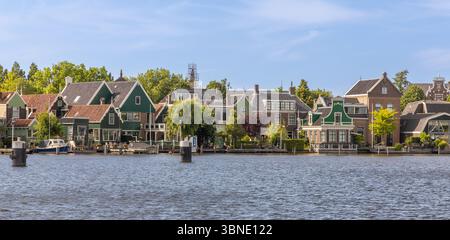 Dieses malerische Foto fängt die traditionellen grün bemalten niederländischen Häuser ein, die das ruhige Wasser von Zaanse Schans säumen, unter einem klaren blauen Himmel, Epitomi Stockfoto