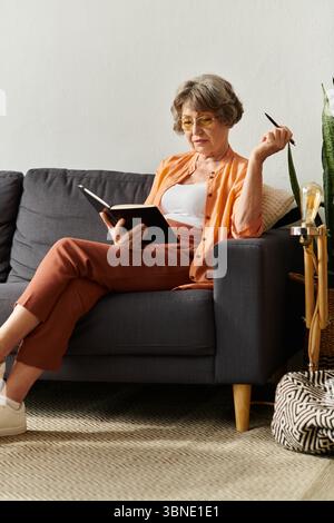 Die reife Frau entspannt sich auf der Couch, liest ein Buch, während sie die gemütliche Atmosphäre des Zuhauses genießt. Stockfoto