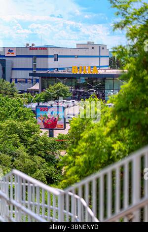 Einkaufszentrum mit BILLA-Schildern, dm-Geschäft, lebendigen Plakatwänden, umgeben von üppig grünem Laub und blauem Himmel. Konzept des Verbrauchermarketings, des städtischen Einzelhandels Stockfoto
