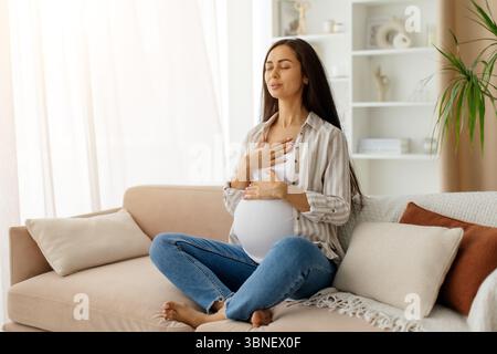 Eine junge schwangere Frau sitzt im Kreuz auf einer Couch und übt tiefe Atmung Stockfoto