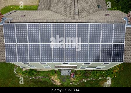Weitwinkelaufnahme einer großen Dachsolarzelle in einem Vorstadthaus, von der Seite aus gesehen mit Schornsteinen, Gartenblumen und einer amerikanischen Flagge. Stockfoto
