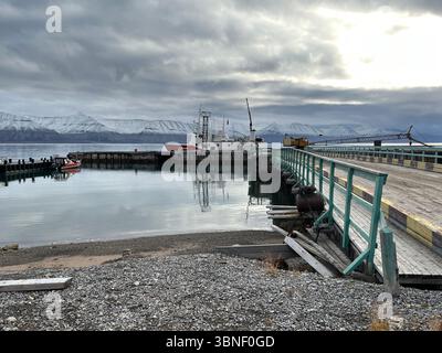 Eine ruhige Küstenszene mit Booten, einem Pier und fernen Bergen unter einem bewölkten Himmel in Pyramiden, Svalbard Stockfoto